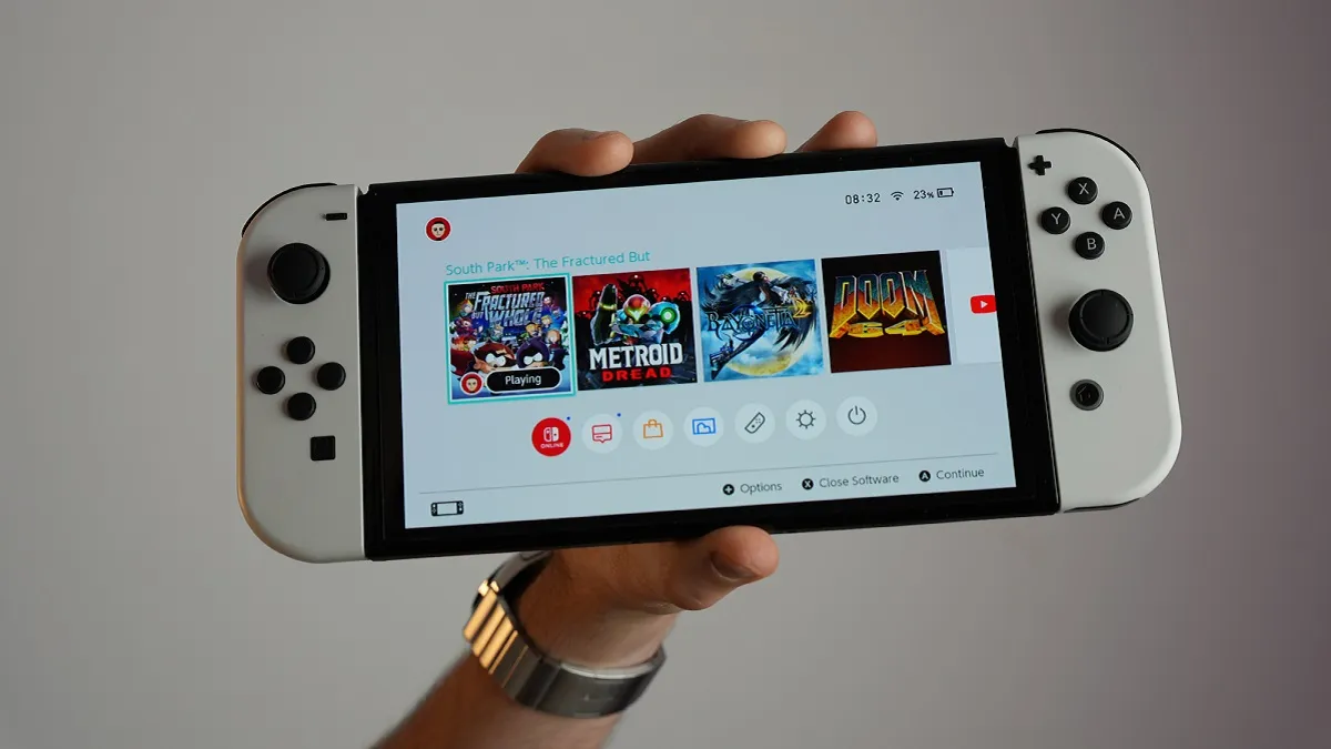 A hand holds up a white Nintendo Switch showing games like Doom 64 and Metroid Dread on the screen.